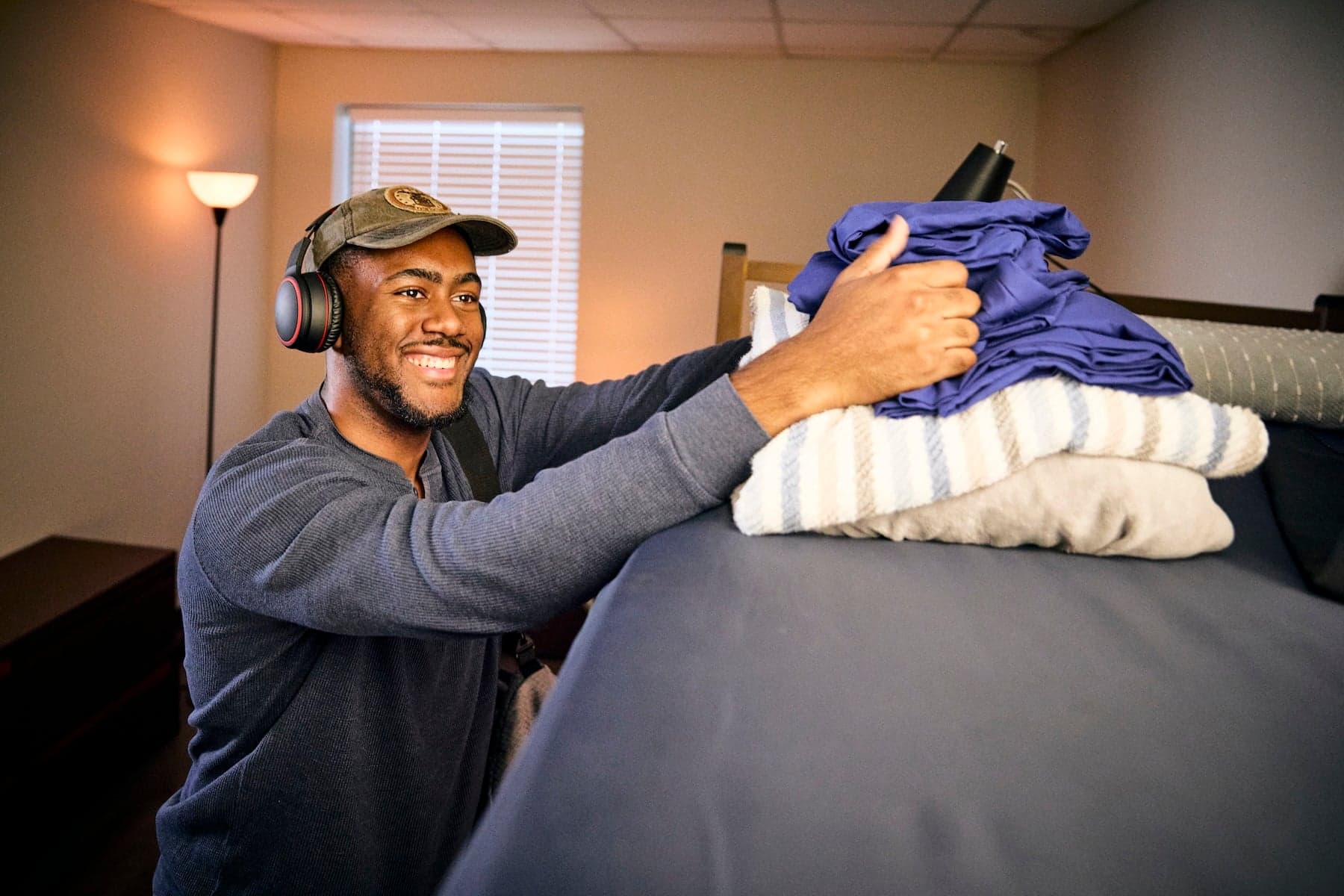 A person wearing headphones and a cap smiles while organizing folded linens on a bed. The room is softly lit, creating a relaxed and cozy atmosphere.