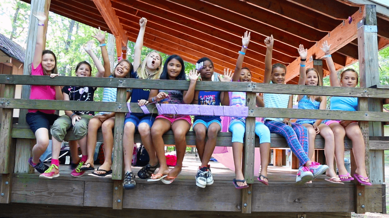 A group of children sit on a wooden bridge, smiling and raising their arms, under a covered structure on a sunny day.