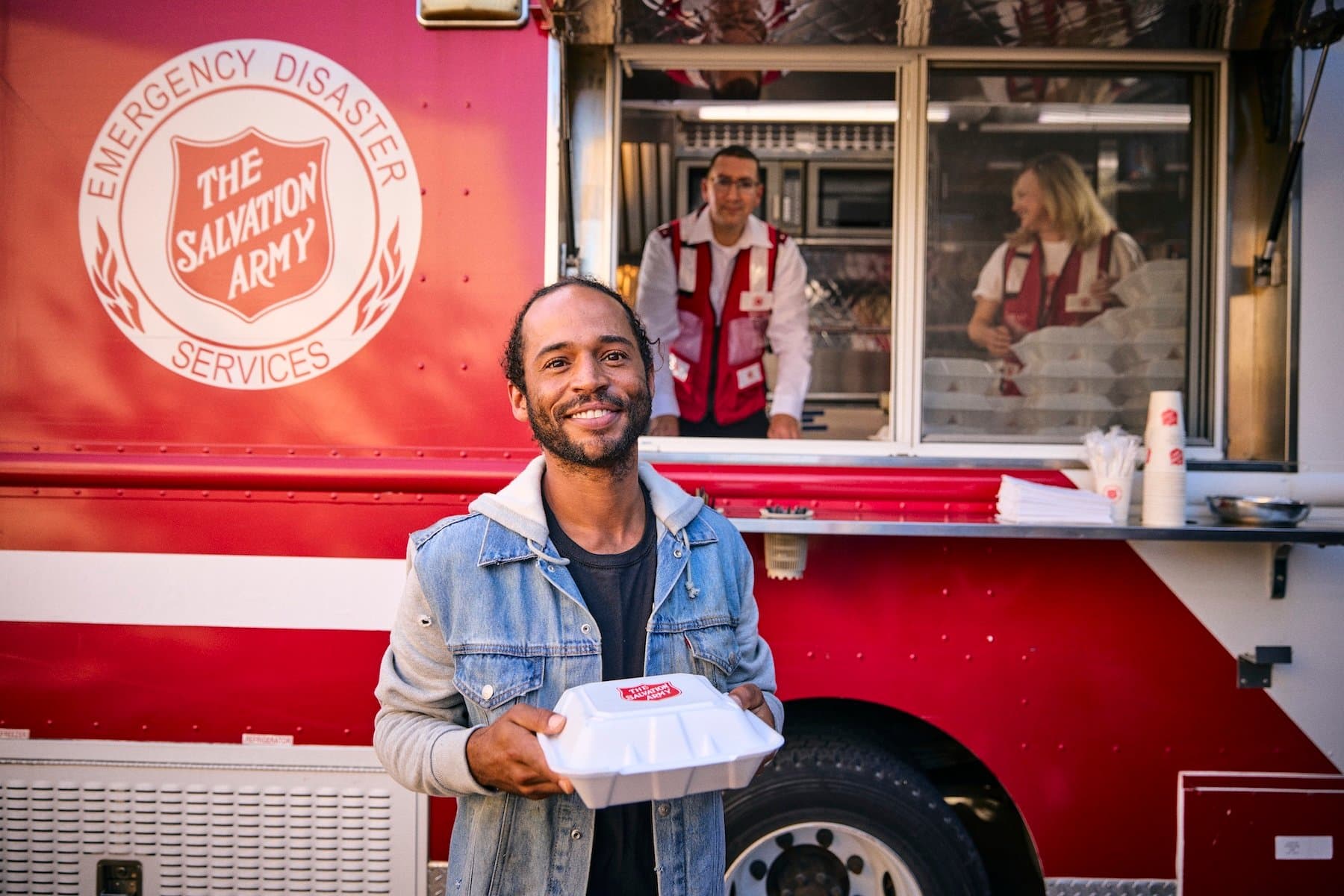 A man smiles while holding a food container in front of a Salvation Army emergency services truck. Two volunteers are inside, ready to serve meals.