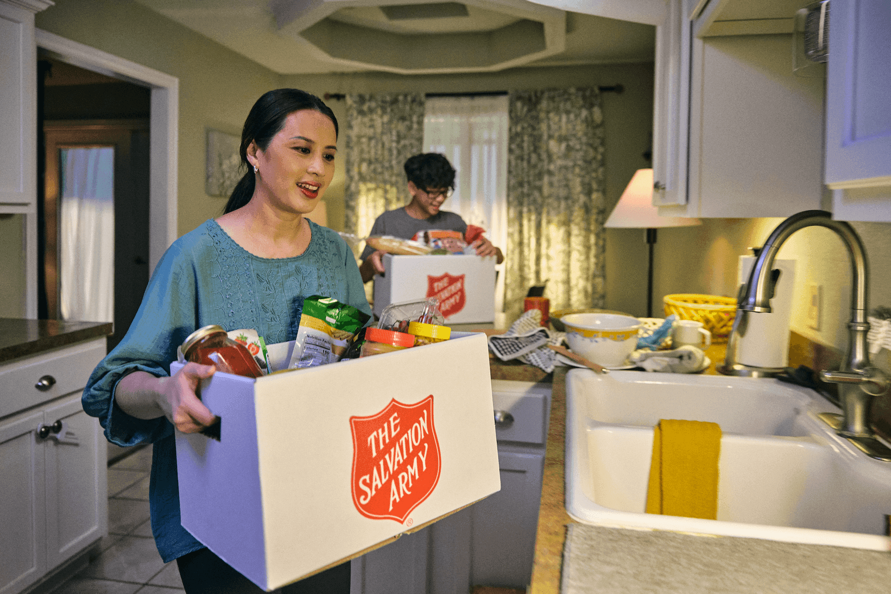 A woman and a child in a kitchen hold Salvation Army boxes filled with food items, smiling. The setting is warm and homey, suggesting a spirit of giving.